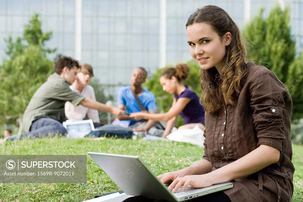 Young woman using laptop computer on grass, people in background