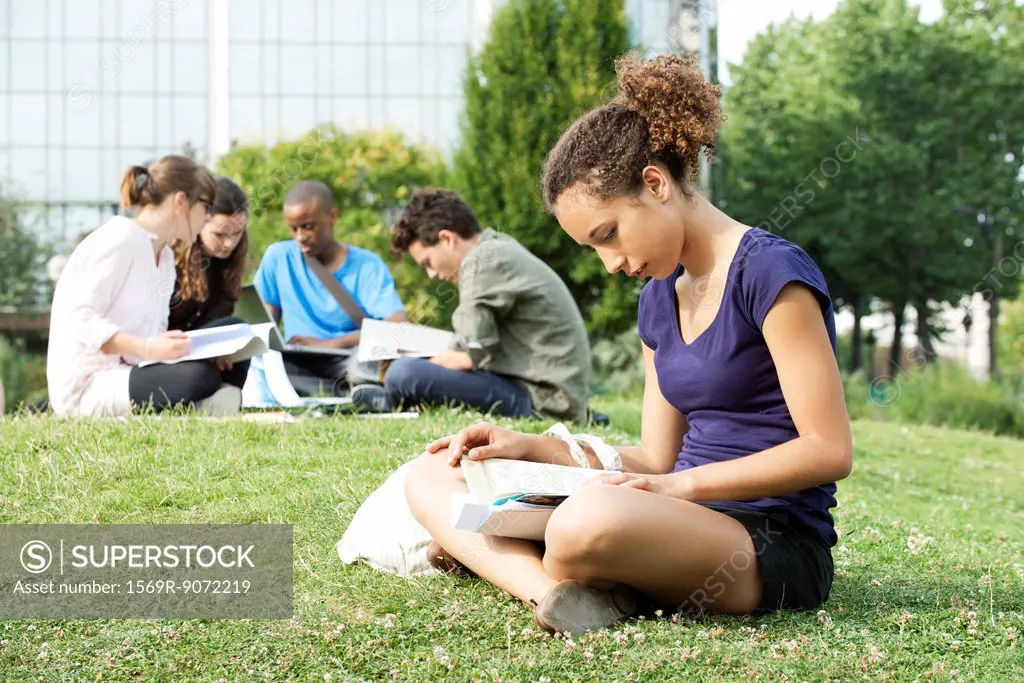 Young woman reading book on grass, group of young people in background