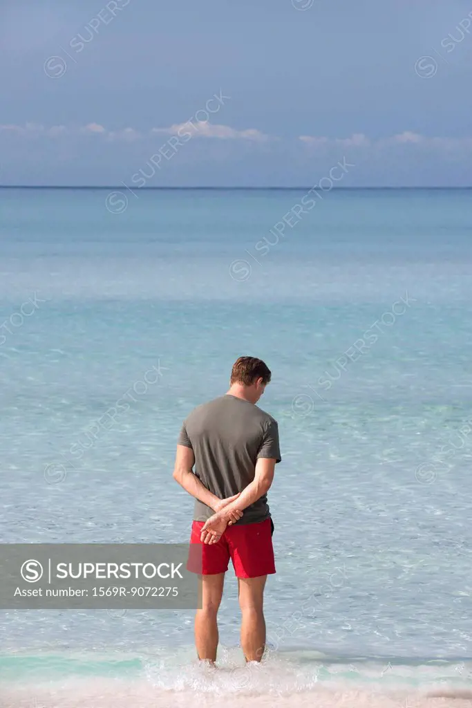 Man standing ankle deep in water contemplating ocean, rear view