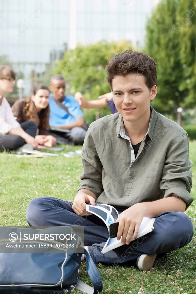 Young man sitting on grass, people in background, portrait
