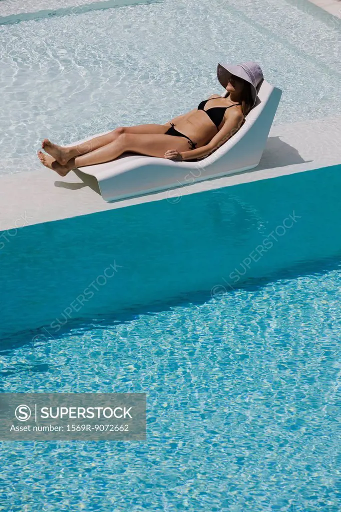 Young woman wearing sun hat reclining on poolside deckchair