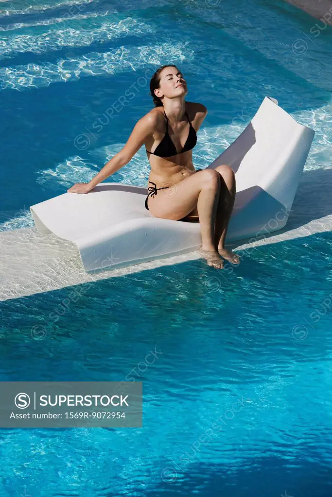 Young woman sitting on poolside deckchair dangling feet into water