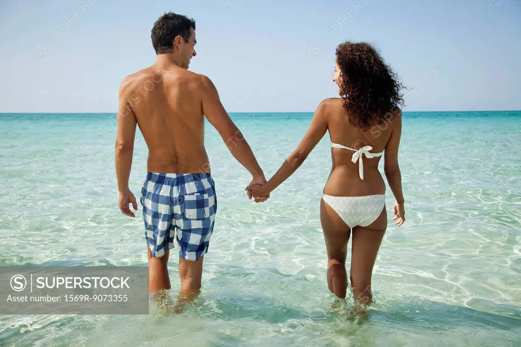 Couple holding hands while wading in sea, rear view