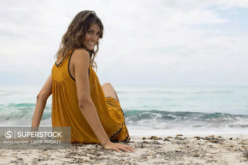 Young woman sitting on beach, looking over shoulder at camera