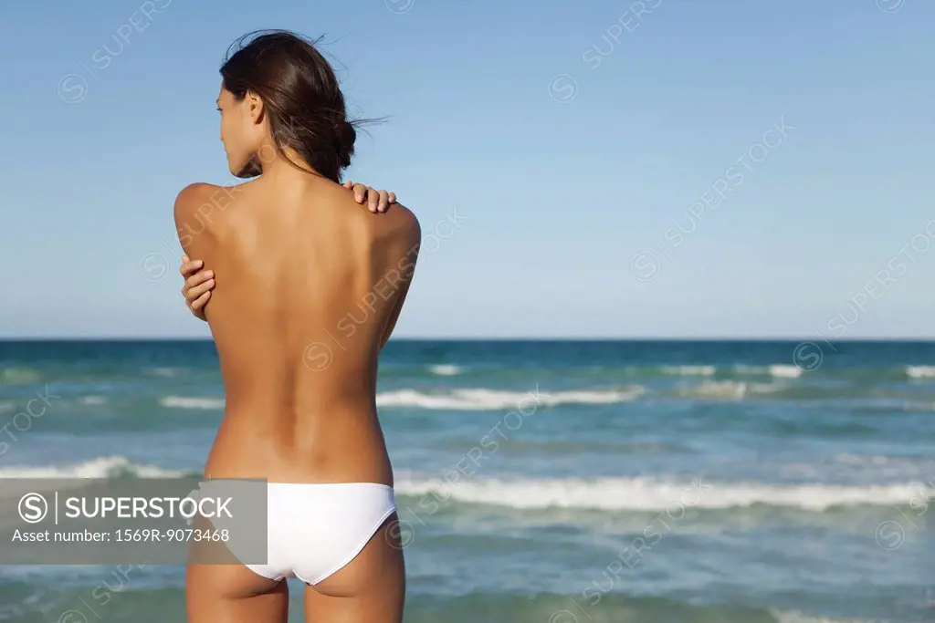 Semi_naked woman in bikini bottom hugging self by ocean, rear view
