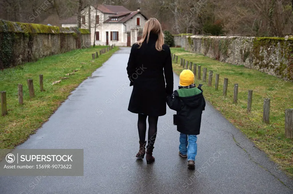 Mother and son walking on path in village, holding hands, rear view