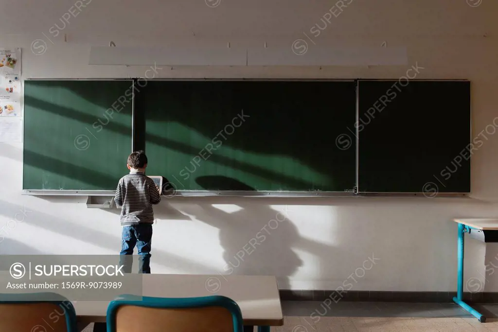 Boy standing in classroom, in front of blackboard, rear view