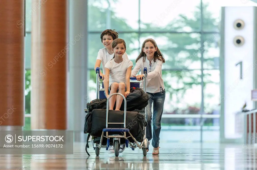 Brother and sister pushing younger sister on luggage cart in airport
