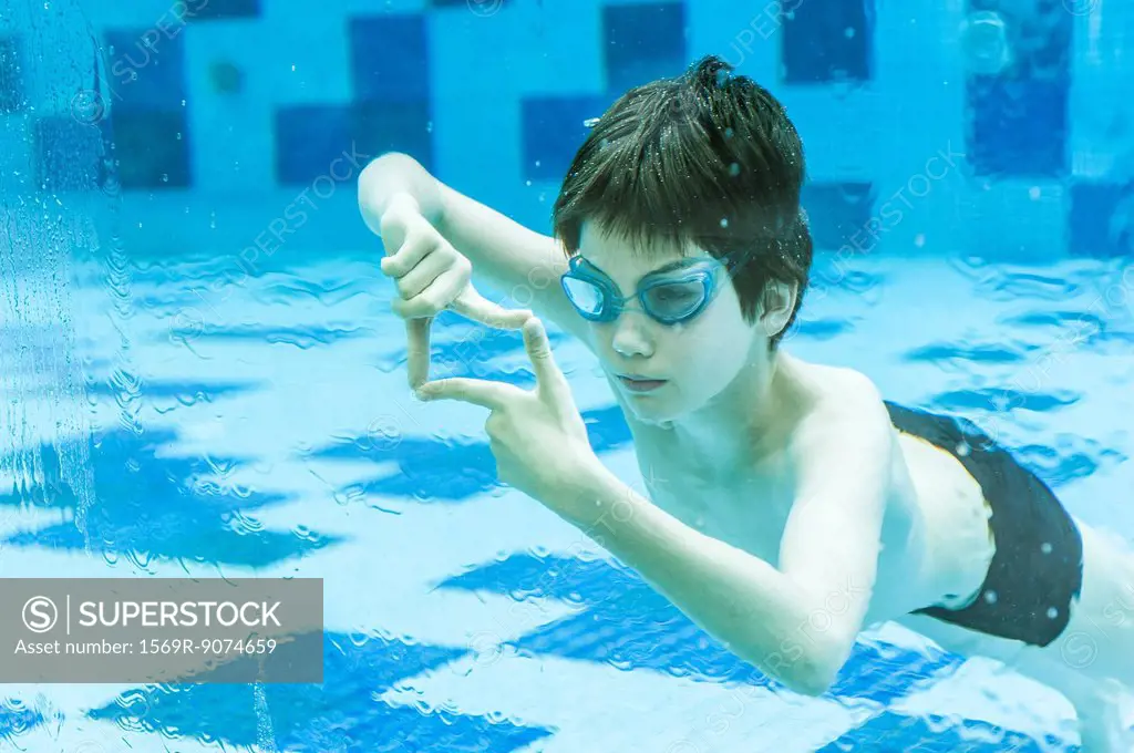 Boy wearing goggles swimming underwater in swimming pool, hands forming finger frame