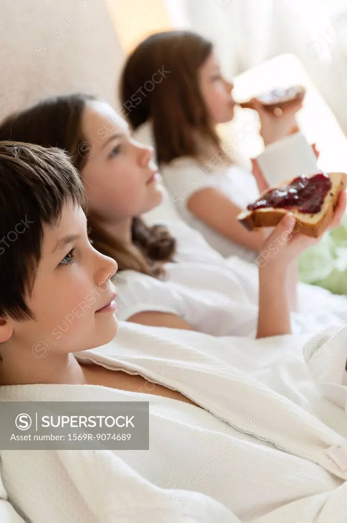 Siblings relaxing on bed, girls eating toasts in background