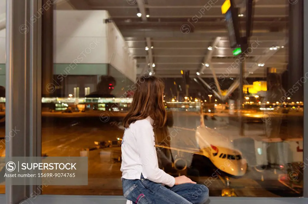 Young girl watches from airport terminal window as flight crew prepare commercial airplane for boarding