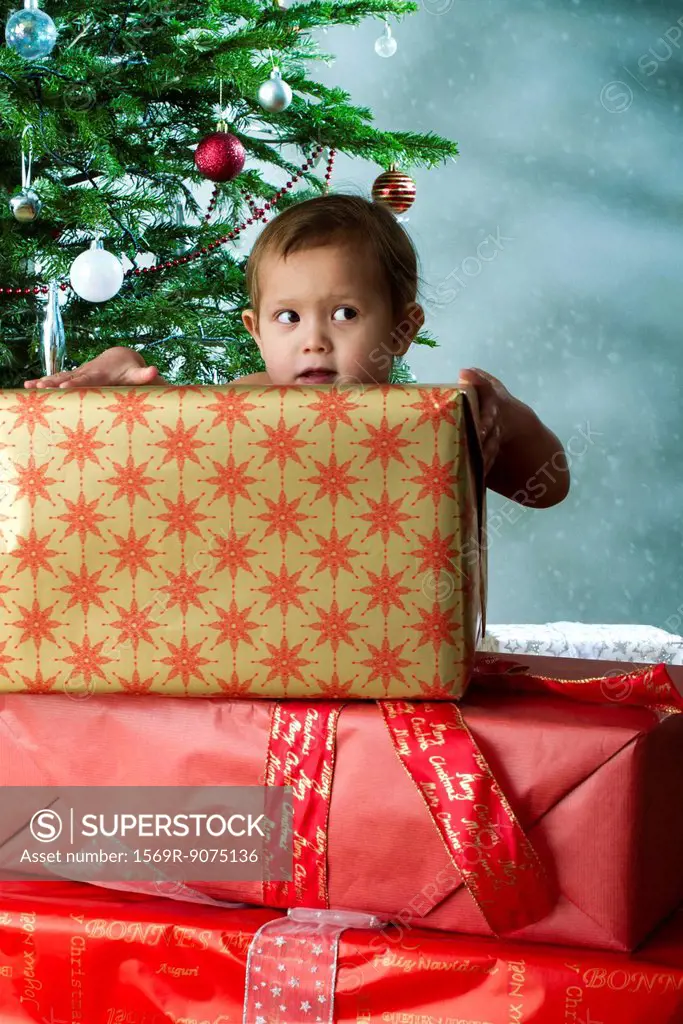 Baby girl standing behind stack of Christmas gifts with mischievous expression