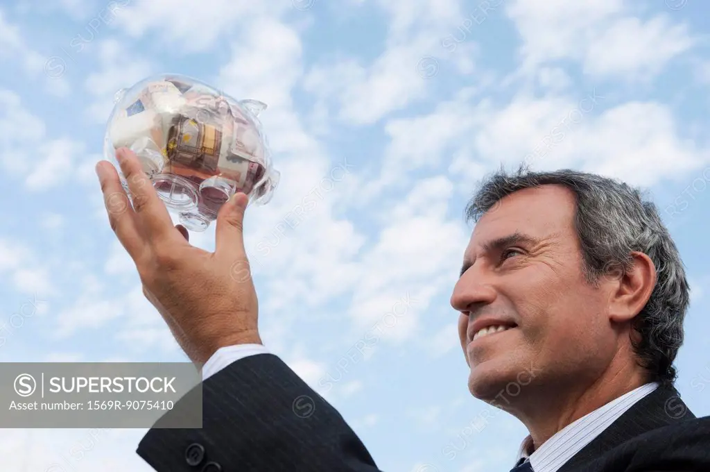 Mature man holding up transparent piggy bank filled with euros