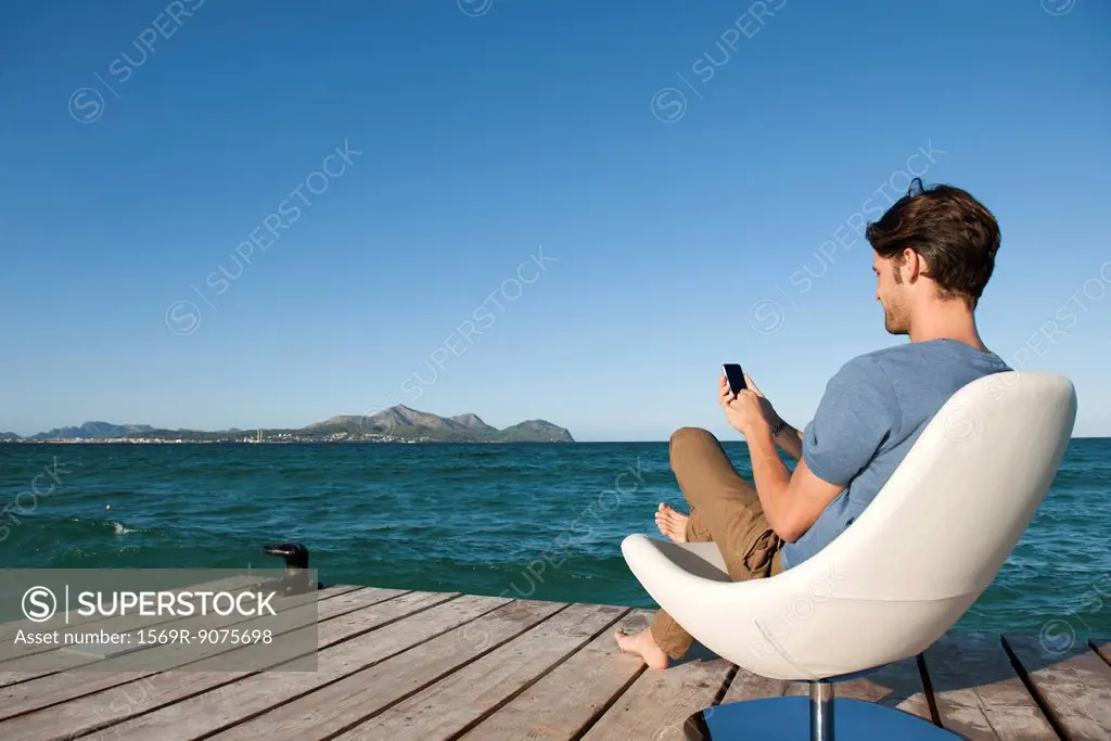 Young man sitting in armchair by lake using cell phone, side view