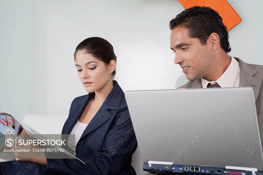 Businessman with laptop computer looking over while businesswoman reads magazine