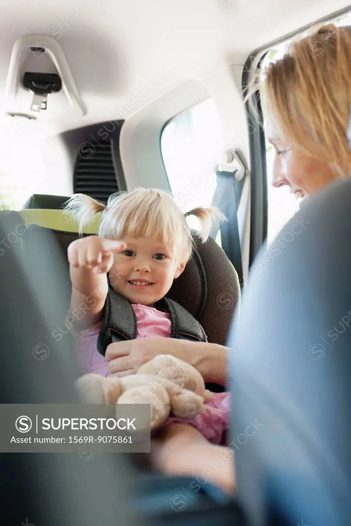 Little girl pointing and smiling as her mother fastens her into car seat