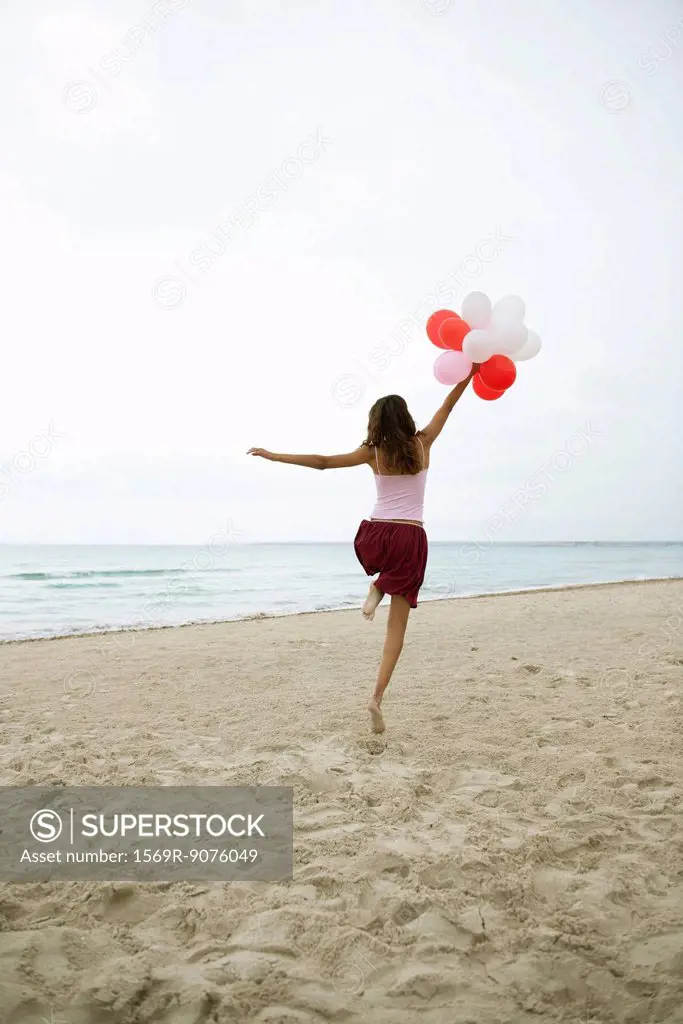Woman running on beach with bunch of balloons, rear view