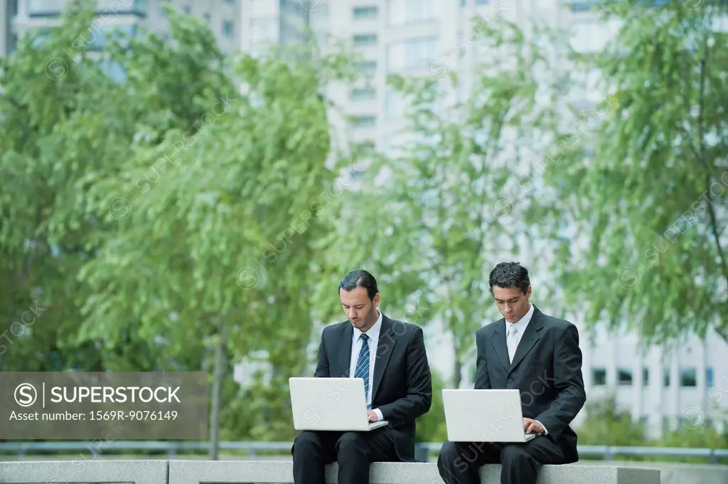 Businessmen sitting side by side using laptop computers outdoors