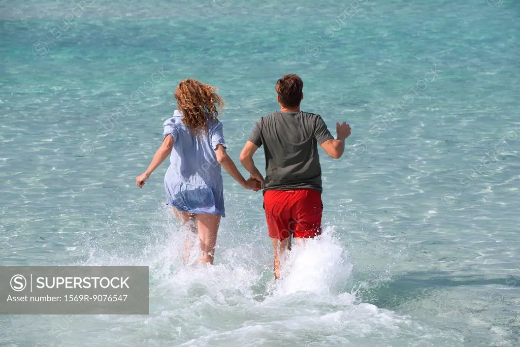 Couple running into water holding hands, rear view