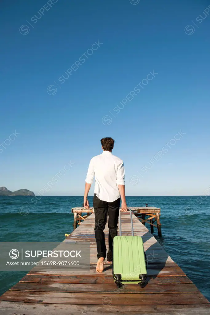 Man pulling luggage walking towards end of pier, rear view