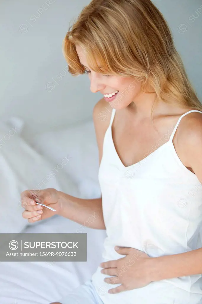 Young woman holding pregnancy test with hand on abdomen, smiling