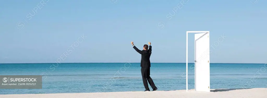 Businessman standing on beach looking at ocean with arms raised, rear view