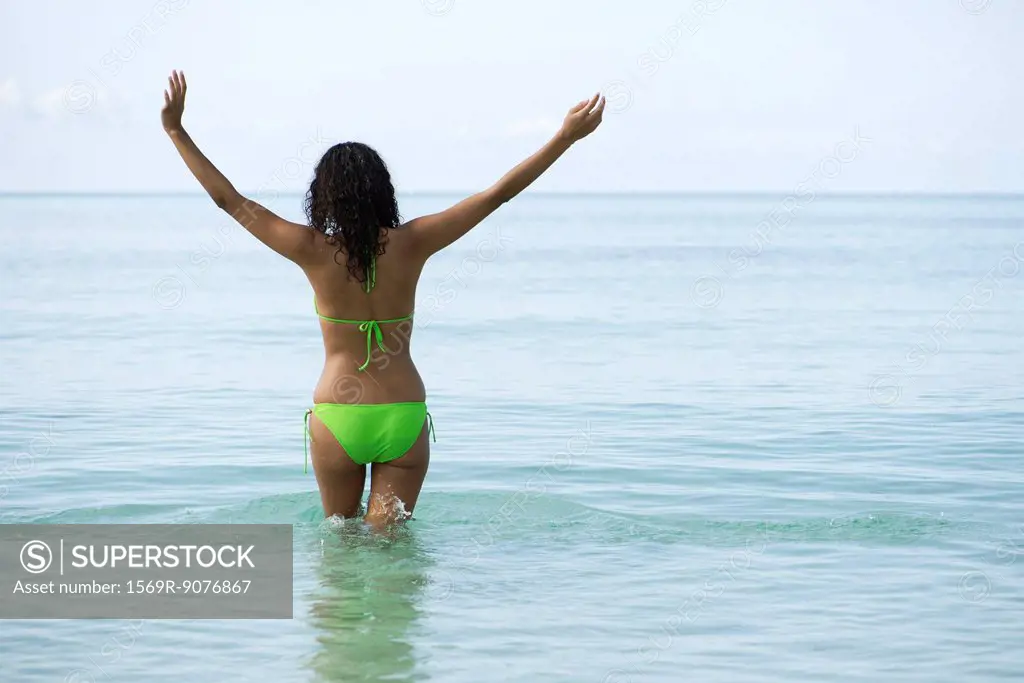 Woman in bikini standing in ocean with arms out, rear view