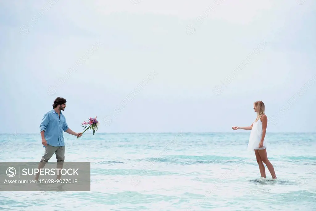 Couple walking in sea towards each other, man holding out bouquet