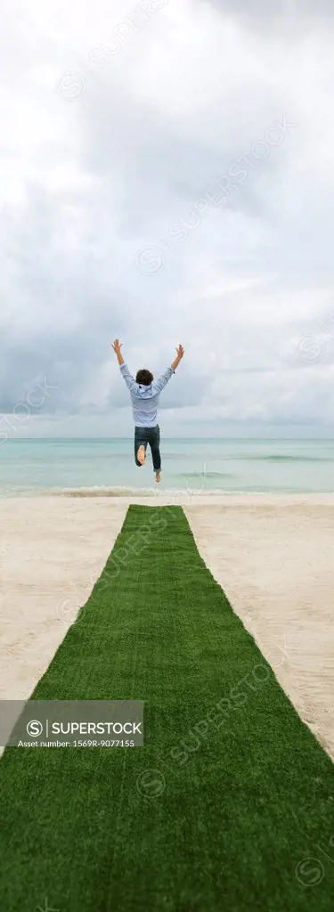Man above artificial turf on beach, arms outstretched, rear view