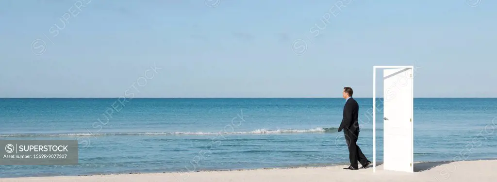 Businessman standing on beach next to half_open door, looking at ocean