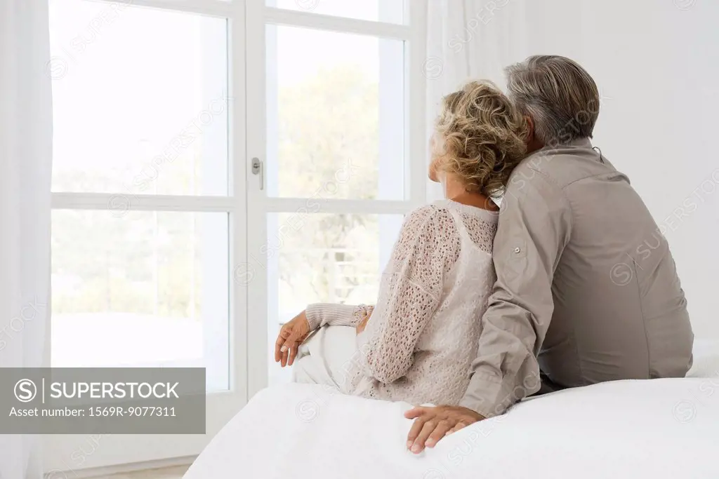Couple sitting together on bed, looking out window, rear view
