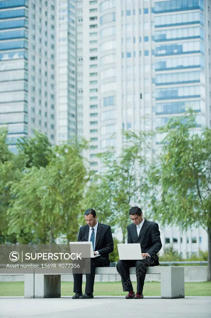 Businessmen sitting side by side using laptop computers outdoors