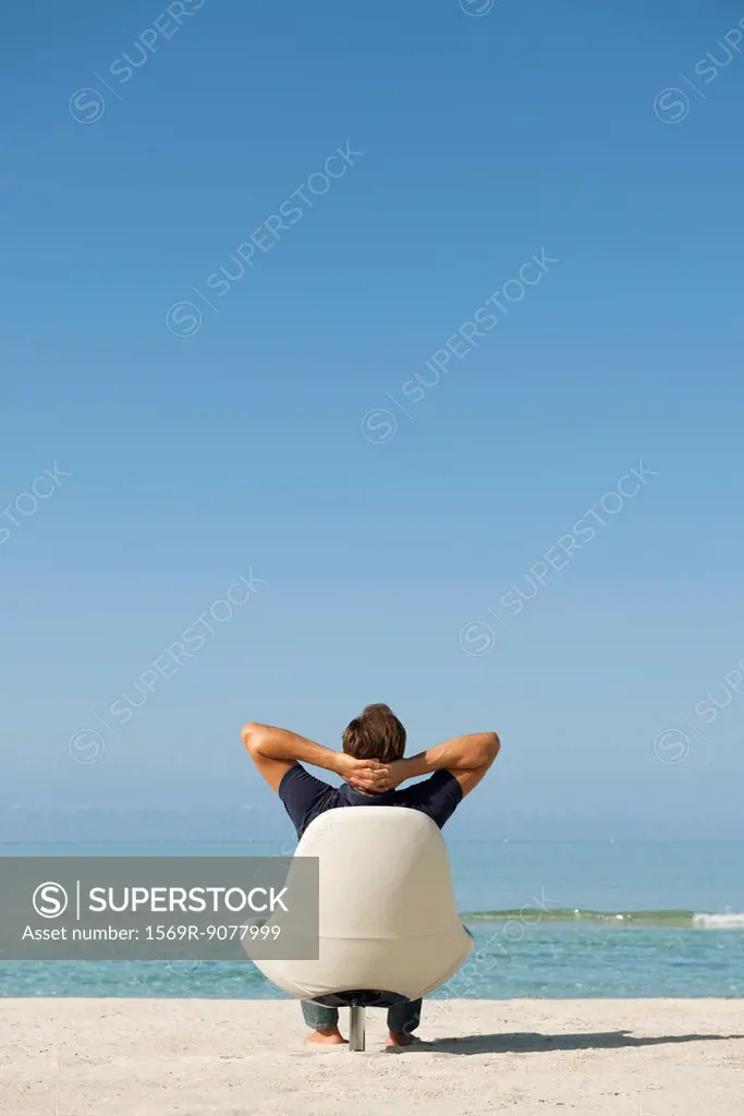 Man sitting in armchair on beach looking at ocean, rear view