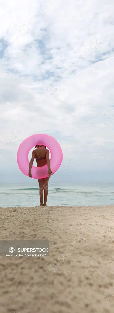 Girl standing on beach with inflatable ring, looking at sea, rear view