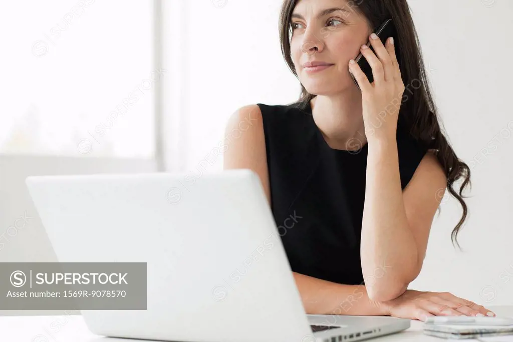 Mid_adult woman sitting at desk with laptop, talking on cell phone