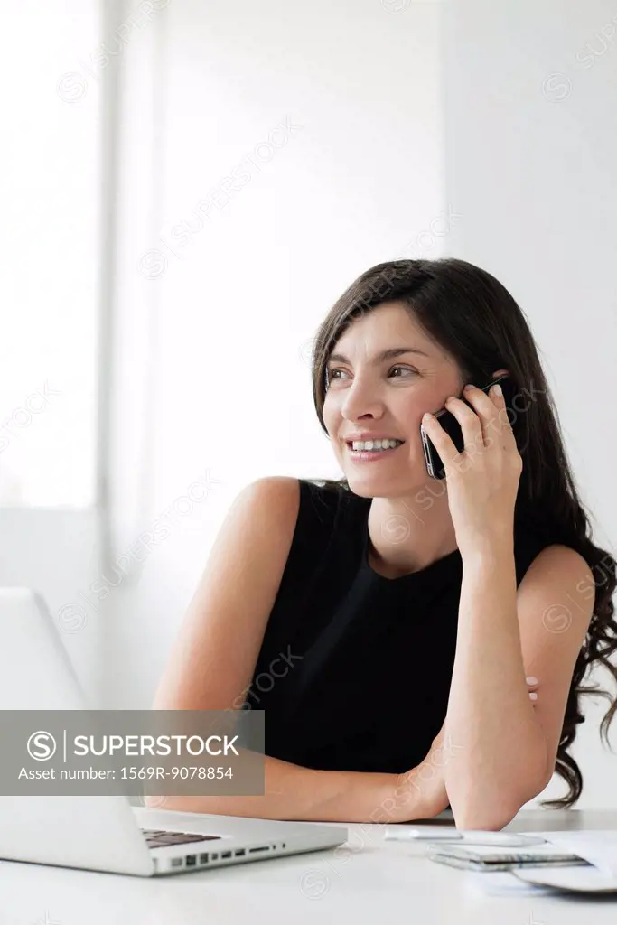 Mid_adult woman sitting at desk with laptop, talking on cell phone