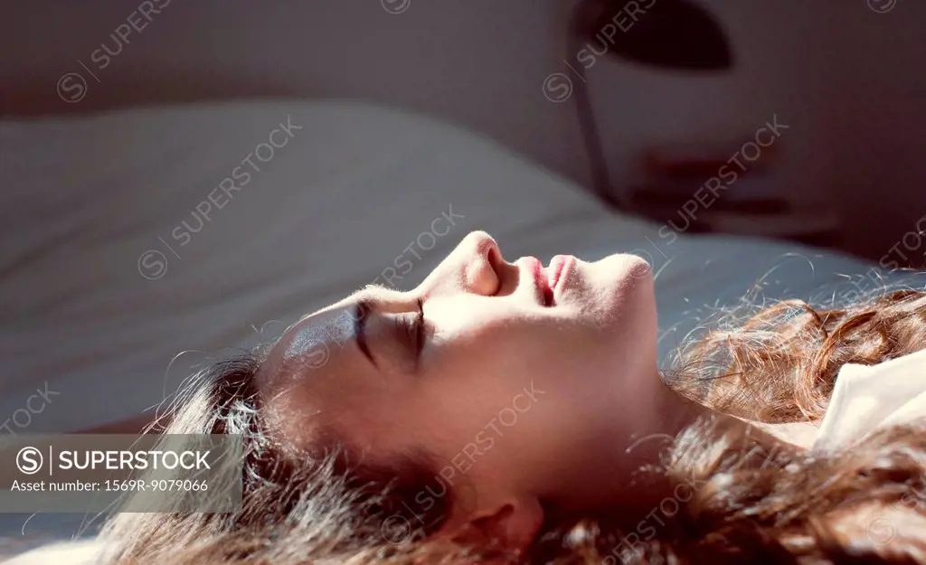 Mid_adult woman lying on bed, enjoying sunlight with eyes closed