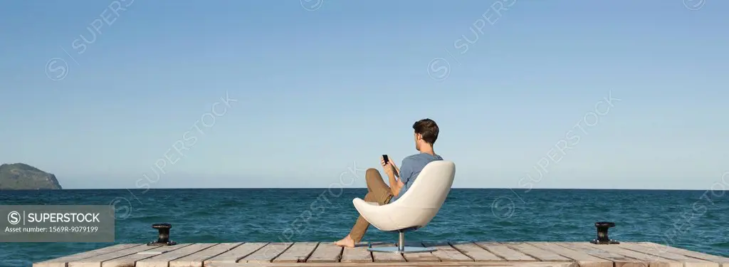 Man siitng in chair on dock using cell phone, rear view