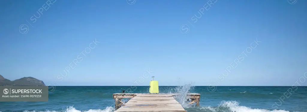 Waves crashing against dock, yellow suitcase at end of dock