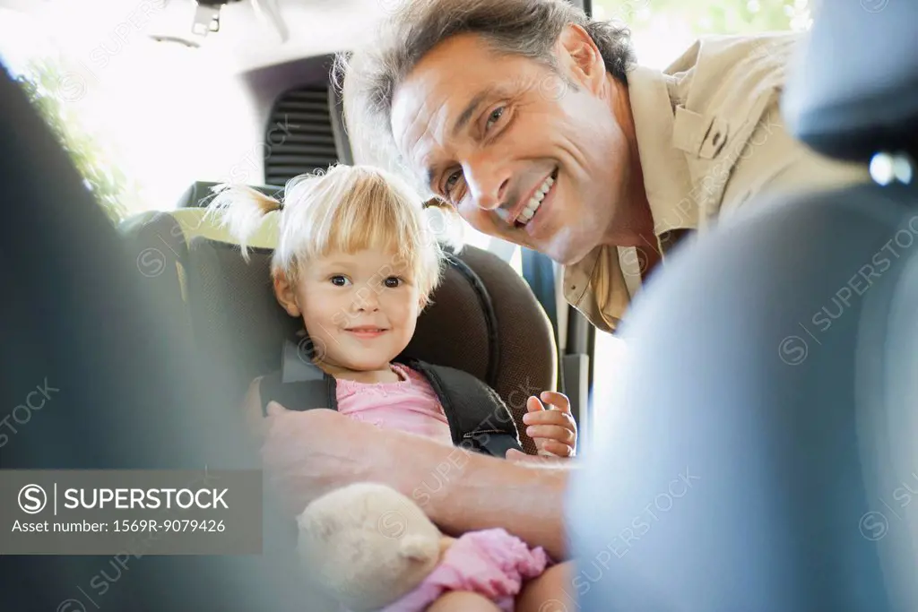 Father fastening little girl into car seat, both smiling at camera