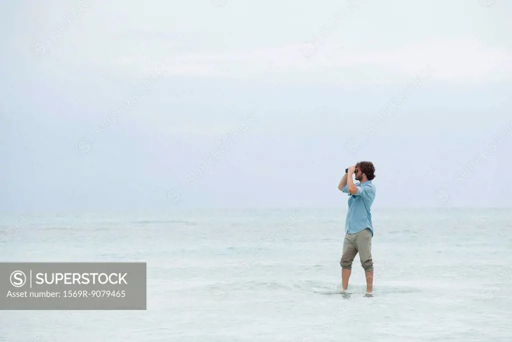 Man standing in sea, looking through binoculars