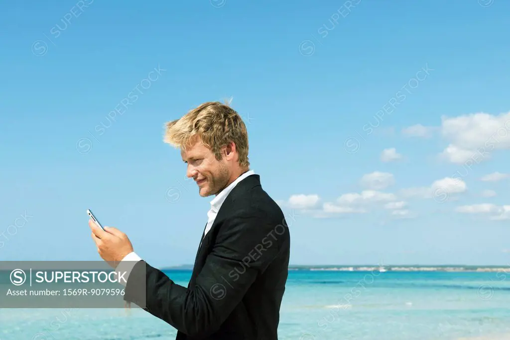 Young businessman using cell phone by ocean, side view