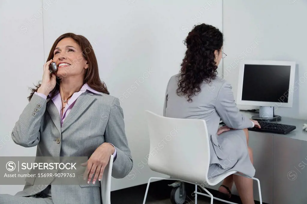 Businesswoman idly chatting on cell phone while colleague works nearby