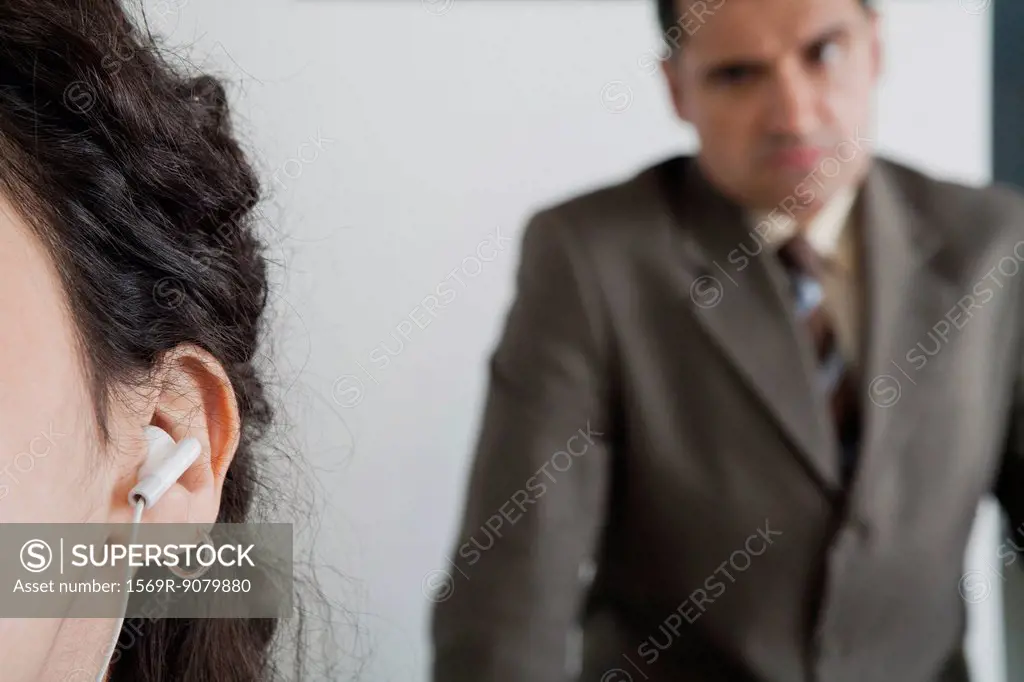 Woman listening to earphones in office, angry boss in background