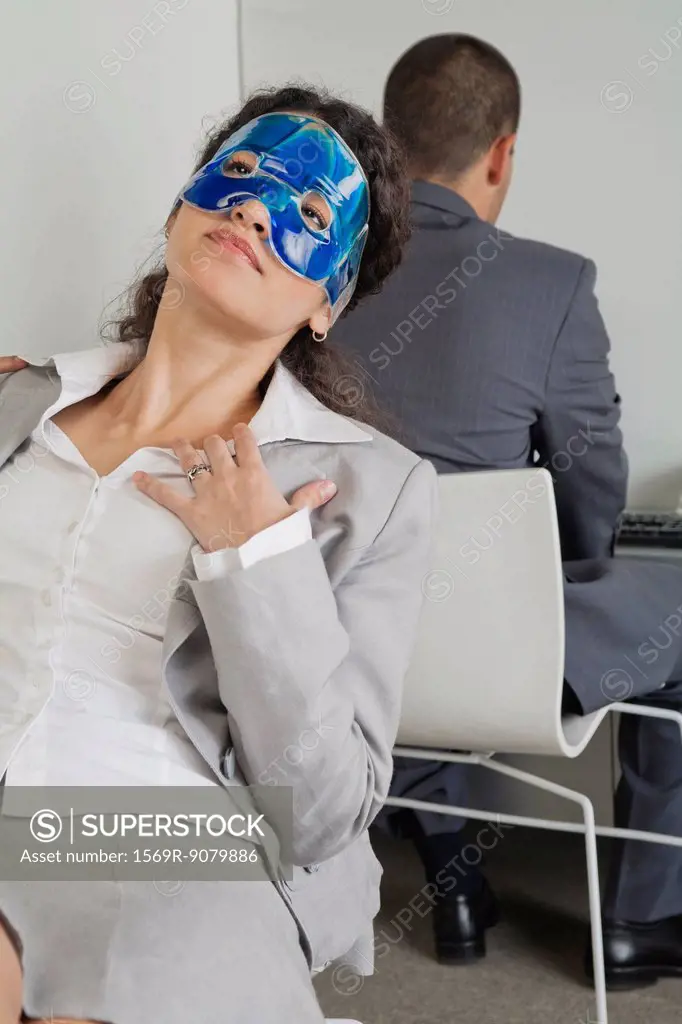 Woman relaxing with eye mask in office as colleague works nearby