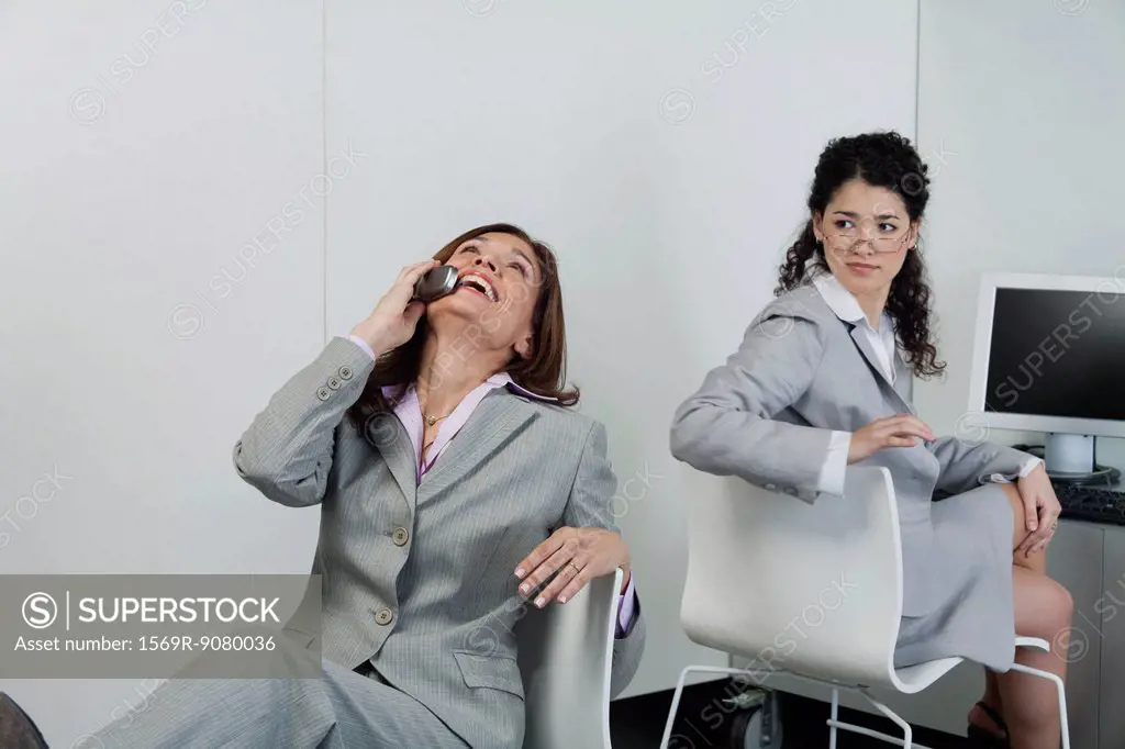 Woman looking over shoulder at coworker laughing on cell phone in office