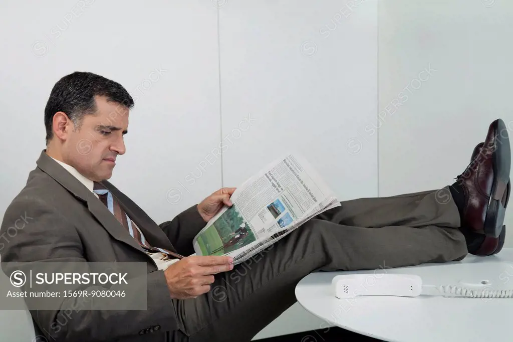 Businessman reading newspaper in office with feet up on desk