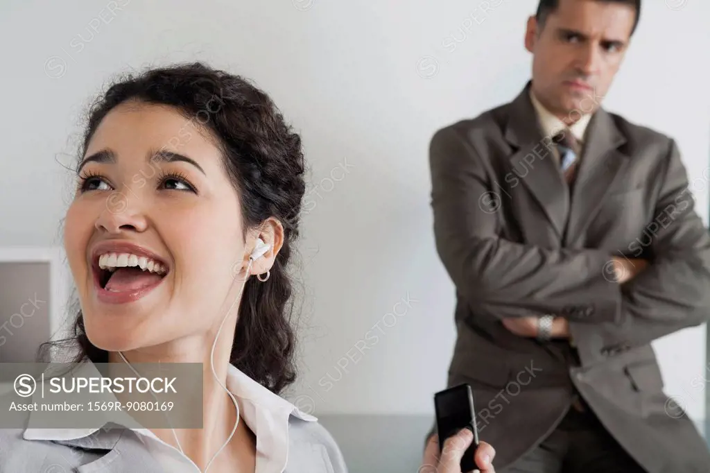 Woman distractedly listening to MP3 player at work, boss glaring in background