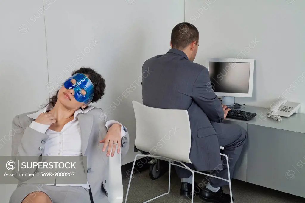 Woman napping with eye mask in office as colleague works nearby