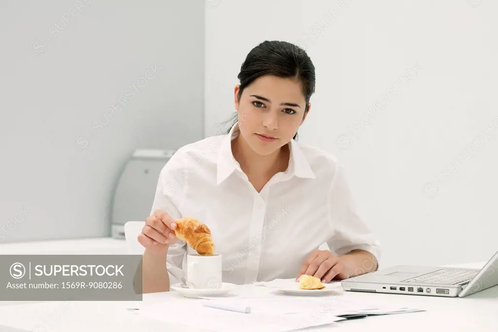 Young woman dipping croissant in coffee at desk in office